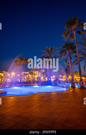 Illuminated resort swimming pool and fountains at night, surrounded by tall palm trees, warm hotel lights and a tiled terrace under a deep blue sky. Stock Photo