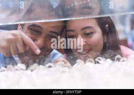 Middle Eastern couple happily browsing wedding rings behind a glass display while friends and family look on, sharing excitement and anticipation duri Stock Photo