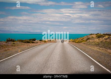 A straight two-lane road stretches toward the vivid turquoise waters of Lago Argentino under a partly cloudy Patagonian sky. Stock Photo