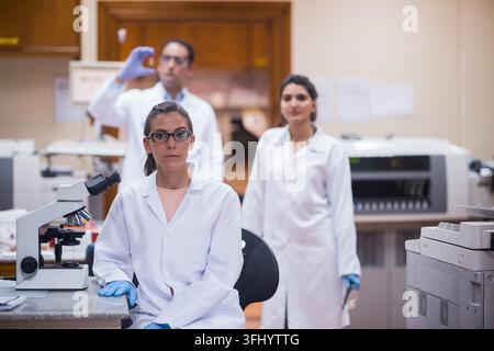 Middle Eastern and Arab young doctors and a lab technician in white coats perform blood testing and diagnostic analysis, using a microscope and lab eq Stock Photo