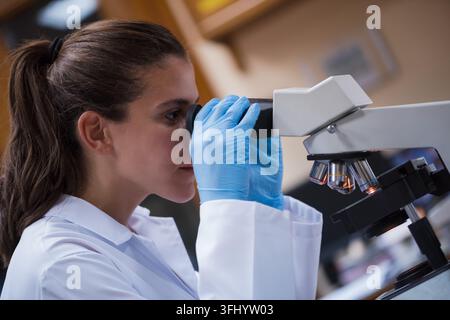 Young medical technician in a white lab coat and blue gloves peers into a microscope while analyzing blood samples, conveying focus, professionalism a Stock Photo