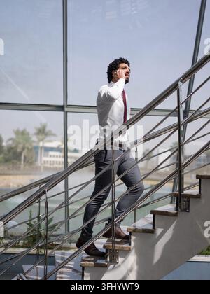 Middle Eastern corporate professional climbs a stairwell in a modern office while talking on his phone, portraying focus, mobility and a busy startup Stock Photo