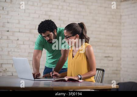 Two young Middle Eastern business professionals collaborate over a laptop and notebook in a bright, creative coworking startup space, discussing ideas Stock Photo