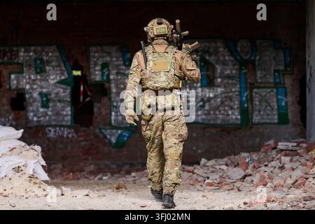 A camouflaged soldier carries a rifle and full tactical gear while walking through a ruined, graffiti-covered urban building during a military trainin Stock Photo