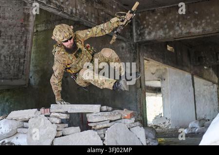A special forces soldier in full camouflage executes a dynamic urban assault jump over rubble while holding a rifle, illustrating tactical movement, c Stock Photo