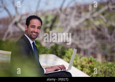 Middle Eastern man in a suit works on a laptop and smiles while seated in a modern outdoor office area, conveying professionalism, confidence and remo Stock Photo