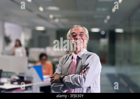 Smiling senior businessman stands with arms crossed in a modern open-plan corporate office, projecting confidence and leadership while colleagues work Stock Photo