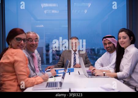 A diverse Middle Eastern and international business team sits around a conference table in a modern glass office, smiling and collaborating on laptops Stock Photo