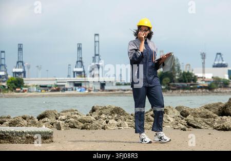 Female maritime engineer using digital tablet for logistics management and industrial port operations near cargo vessel Stock Photo