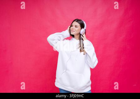 Caucasian Woman in White Hoodie Listening Music with Wireless Headphones on Red Background Stock Photo