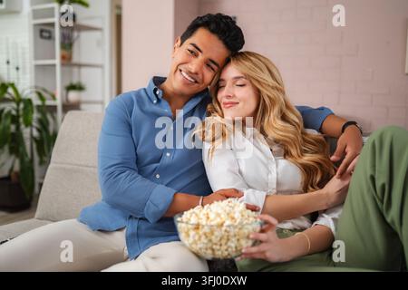 Happy young couple relaxing on sofa watching movie eating popcorn Stock Photo