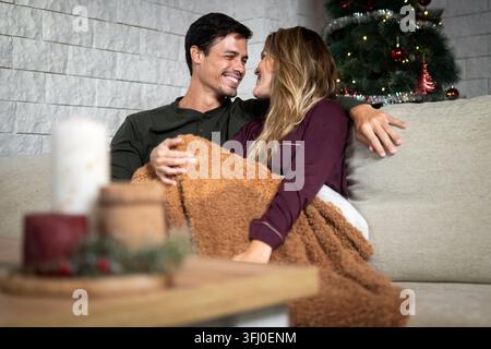 Happy couple sitting close together under a warm blanket and sharing an affectionate moment on the sofa with a decorated Christmas tree in the backgro Stock Photo