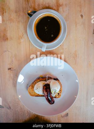 Overhead view of a breakfast plate of poached eggs and a grilled sausage on toast, served with a cup of black coffee on a wooden table. Stock Photo