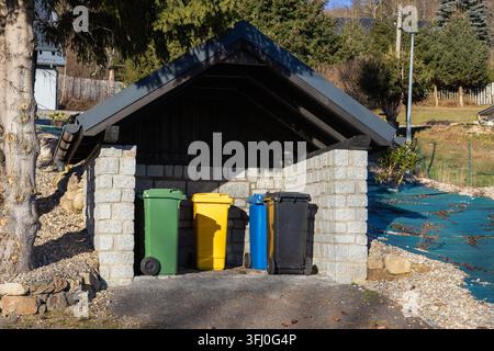 Multicolored waste containers standing in a stone shed within a sunny backyard, representing organized household sorting practices for sustainable liv Stock Photo