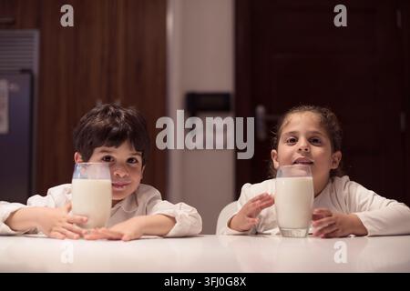 Two Middle Eastern children sit at a kitchen table in a modern home, smiling as they hold glasses of milk during an intimate iftar dinner for Ramadan Stock Photo