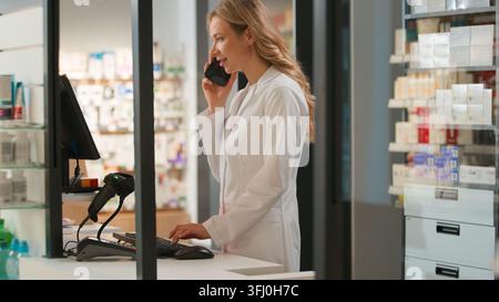 Caucasian woman pharmacist talking mobile phone using computer for online order in pharmacy female medical worker talk smartphone with client patient Stock Photo