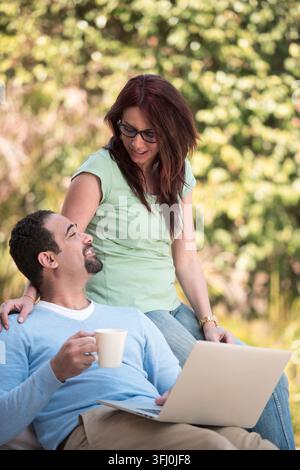 Middle Eastern couple relax outdoors, sharing coffee while using a laptop and smiling together. A warm, casual family moment showing connection, leisu Stock Photo