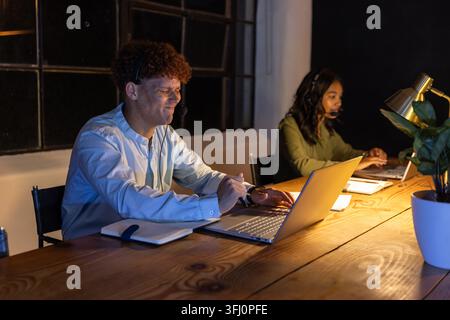 African American coworkers wearing headsets working late at office desk typing on laptops. Collaboration, professionalism, technology, workspace, team Stock Photo