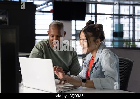 Diverse coworkers working at open-plan office desk, woman in denim jacket typing on silver laptop. Collaboration, teamwork, modern, bright, workspace, Stock Photo
