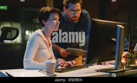 Business team partners with computer discuss work at night evening office. Caucasian man leader coming checking working process help with difficult Stock Photo
