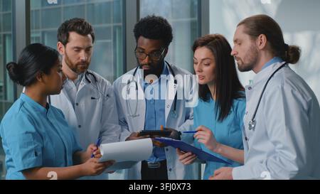 Focused multiethnic five doctors with digital tablet working talking deciding medicine diagnosis in clinic hallway. Multiracial nurses practitioners Stock Photo