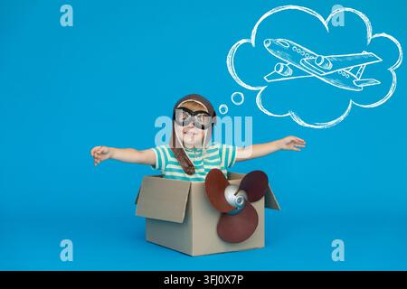 Happy child dreams of becoming a pilot. Kid having fun against blue paper background. Boy wearing striped shirt playing in cardboard box. Summer vacat Stock Photo