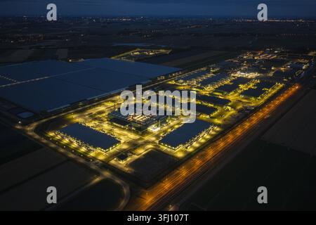 Aerial view of a large data center complex illuminated at night, featuring rows of industrial buildings, cooling systems, and surrounding roads. Stock Photo