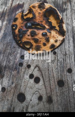 Burnt round flatbread with dark charred spots on a weathered wooden table, shown from above with scattered black crumbs. Stock Photo