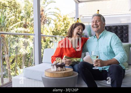 Senior couple celebrating on balcony, red blouse, blue shirt, holding wine and gift, cake with 50. Elderly, duo, terrace, patio, sunny, tropical, fest Stock Photo