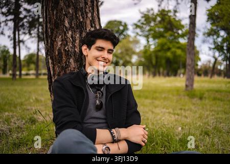 Young man enjoying nature, leaning on tree trunk in park with headphones Stock Photo