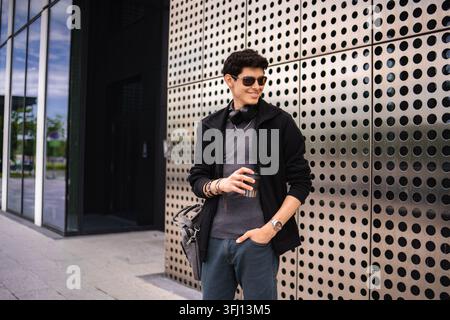 Young man smiling, holding coffee, standing near modern building Stock Photo