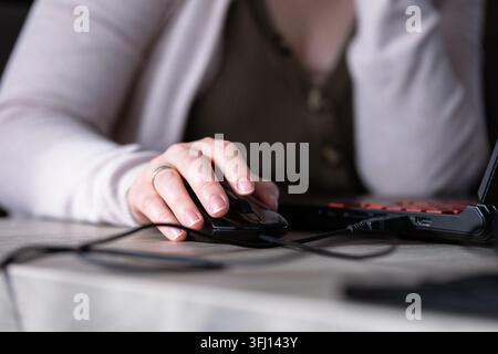 Close-up of a woman hand using a wired black computer mouse on a wooden desk next to a laptop. Natural lighting, soft focus background, calm and focus Stock Photo
