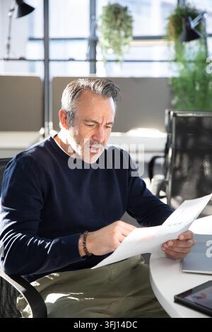 Middle-aged man holding printed sheets, pointing and sitting at round table in office with tablet. Sunlit, modern, minimalist, bright, professional, w Stock Photo