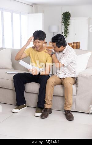 Asian father and son sitting on light-gray couch in living room, holding white envelope and tablet. Letter, family, lifestyle, minimalist, modern, sto Stock Photo