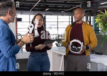 Diverse coworkers discussing VR headset component while holding tablet and full headset at office. Collaboration, prototype, device, hardware, dayligh Stock Photo