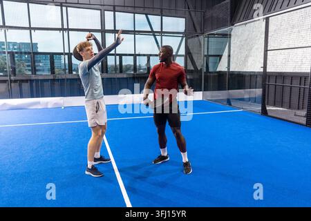 Diverse male sports partners playing padel with rackets on blue court inside glass walled facility. Athletes, teamwork, modern, fitness, active, sport Stock Photo