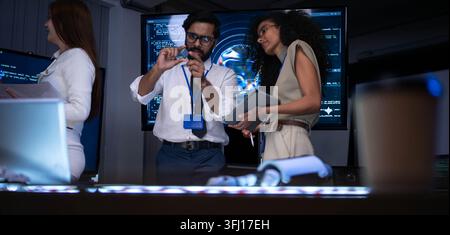 Technology specialists examining electronic component and discussing software engineering solutions in futuristic office with coding screens and digit Stock Photo