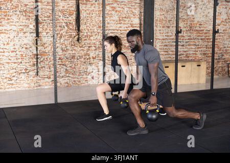 Diverse workout partners in sportswear doing lunges holding yellow kettlebells at gym with rings. Athlete, training, functional, Stock Photo