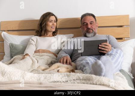Couple sitting on bed, man holding tablet while woman petting dog in pajamas under cream blanket. Duo, bedroom, cozy, neutral, texture, pillows, wood Stock Photo