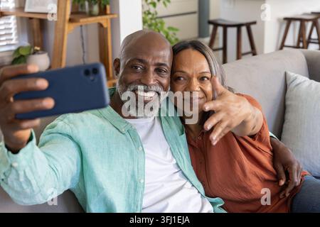 Diverse senior couple sitting on gray sofa at home, smiling and taking selfie with smartphone. Partners, affection, phone, cozy, mature, decor, kitche Stock Photo