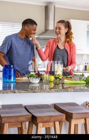 Diverse couple feeding each other while preparing green smoothie in home kitchen with blender. Partners, wellness, blending, cou Stock Photo