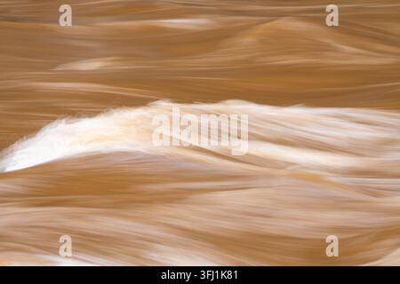 Long exposure captures muddy brown river water flowing in silky smooth waves, creating an abstract pattern of motion and white foam Stock Photo