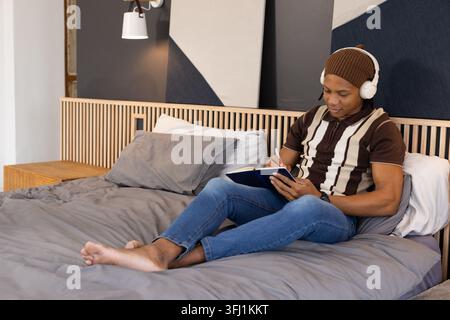 African American man sitting in bedroom writing in notebook with headphones and beanie, copy space Stock Photo