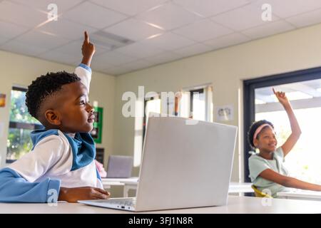 African American school-age students raising hands in bright classroom with silver laptop on desk. Pupils, notebook, technology, education, learning, Stock Photo