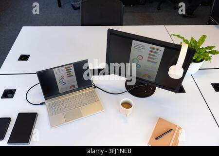 Silver laptop and external monitor are displaying charts on desk, headset hanging, mug sitting Stock Photo