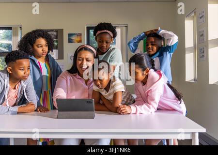 Diverse schoolchildren surrounding adult woman teacher pointing at tablet on classroom table. Device, learning, collaboration, students, instructor, i Stock Photo