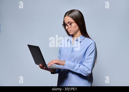Portrait of a focused young woman in glasses and a blue shirt holding an open laptop and typing, standing isolated on a gray background. Concept of re Stock Photo