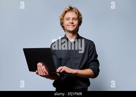 Portrait of a smiling young man holding open laptop computer, typing and looking at camera against light grey studio background. Ideal for e-learning, Stock Photo
