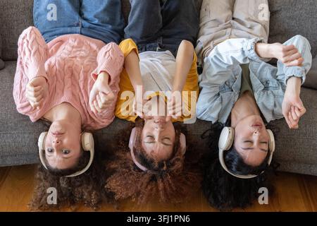 Diverse female friends lying on gray sofa at home wearing over-ear wireless headphones. Trio, lounge, relaxation, music, audio, cozy, casual Stock Photo