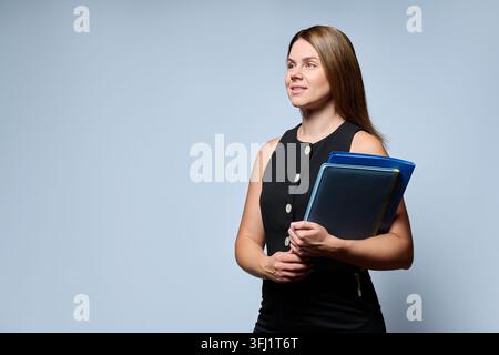 Portrait of a young businesswoman holding document folders and looking away, isolated on gray studio background. Concept of office administration, cor Stock Photo